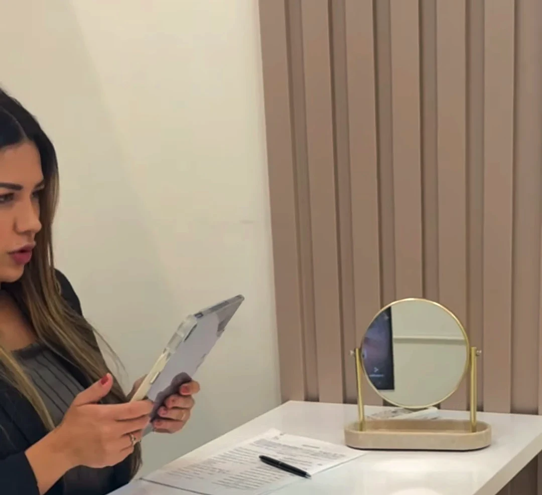 Dra. Daniela Nóbrega à mesa branca, com o tablet nas mãos; sobre a mesa, um espelho redondo dourado e uma ficha com caneta