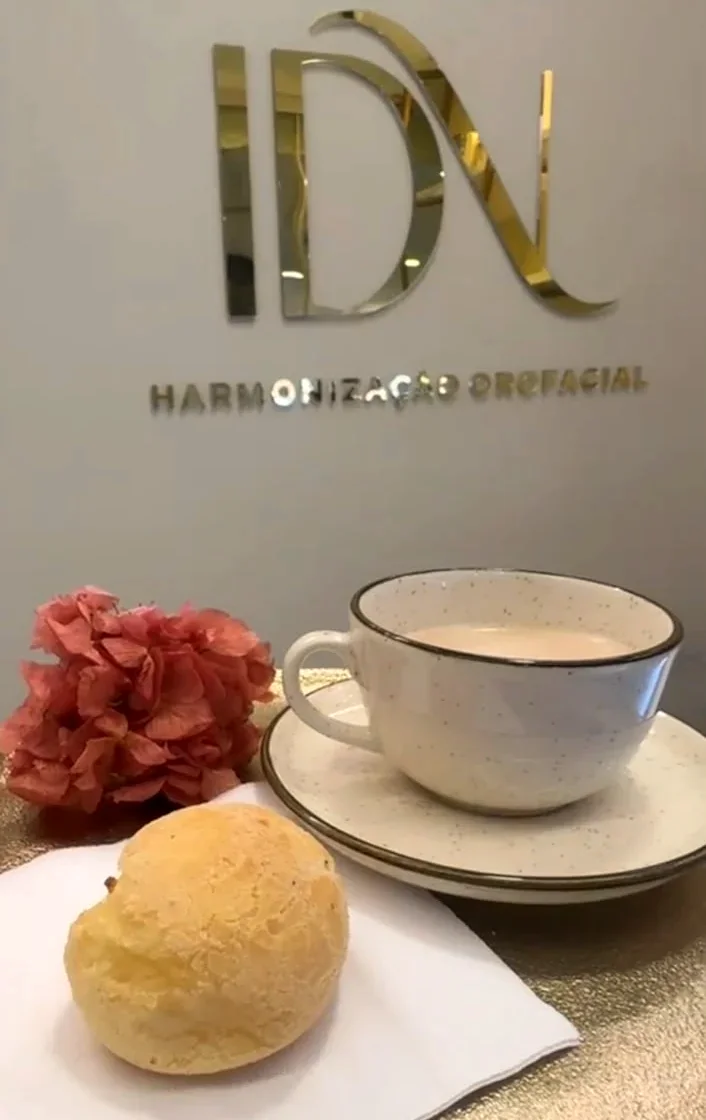 Xícara de café com leite, pão de queijo e uma flor sobre a mesa dourada, diante do monograma IDN na parede.
