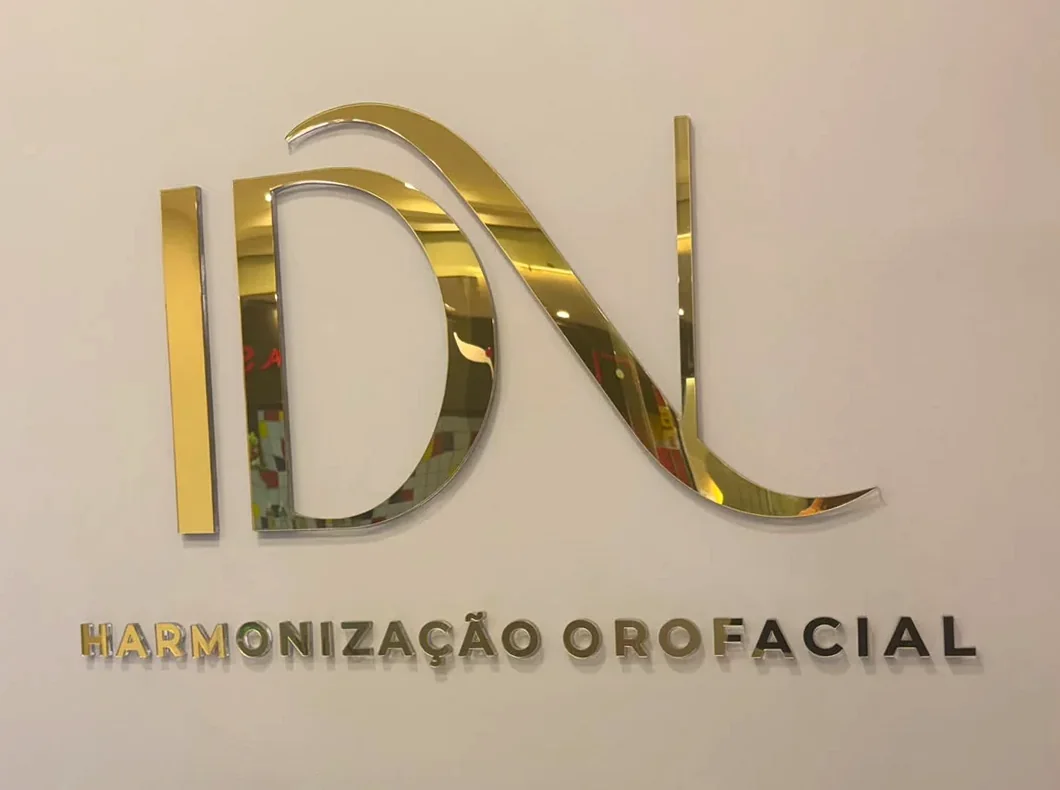 Monograma IDN em acrílico espelhado dourado montado na parede clara da recepção, com as palavras Harmonização Orofacial logo abaixo.