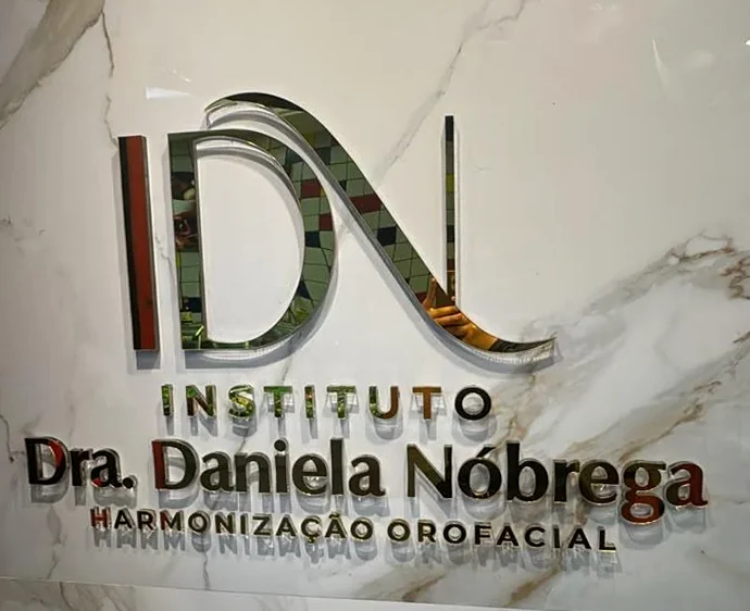 Letreiro espelhado sobre mármore branco de veio caramelo: monograma IDN e, abaixo, Instituto Dra. Daniela Nóbrega, Harmonização Orofacial.