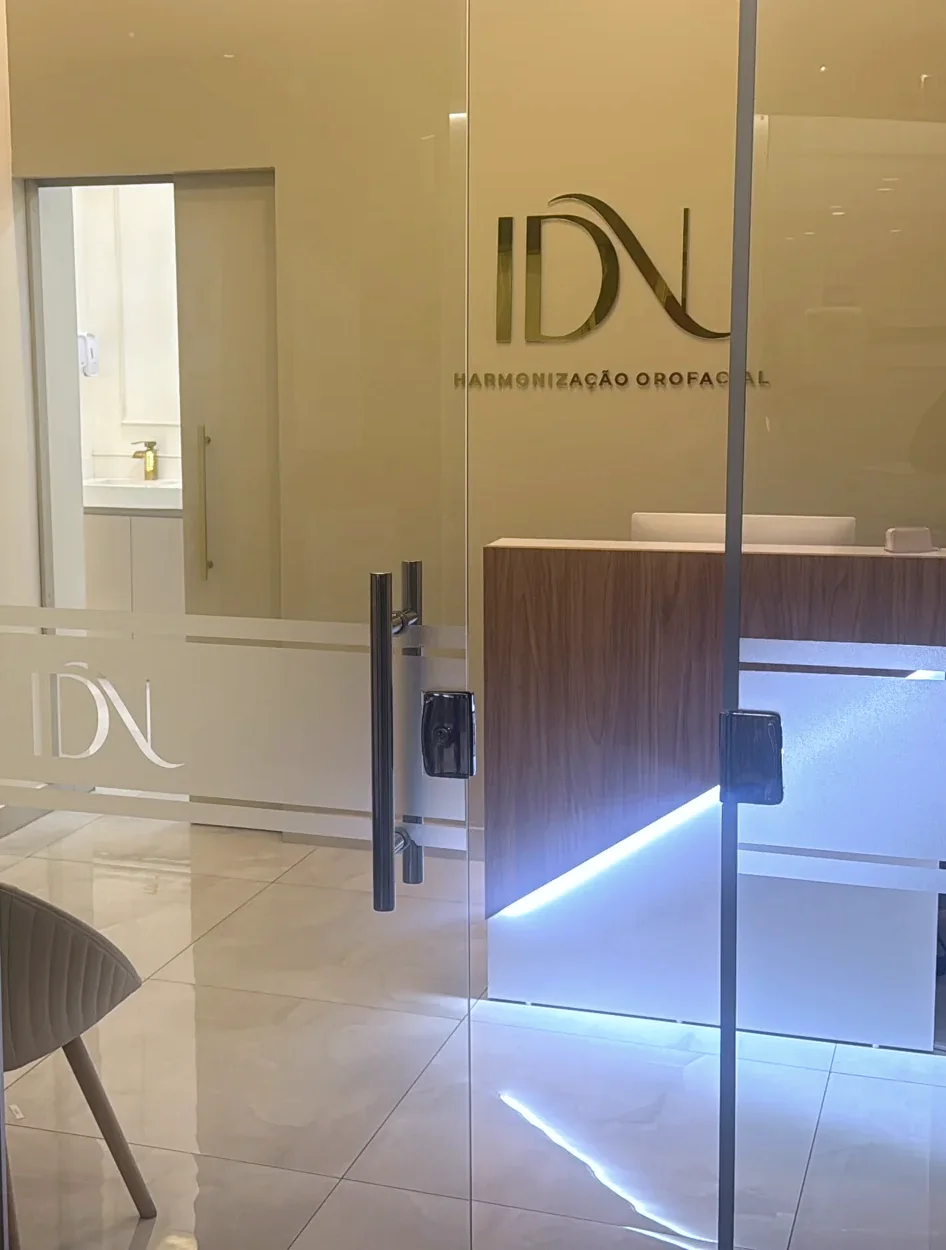 Recepção vista pela porta de vidro: parede clara com o monograma IDN dourado, balcão de nogueira com luz por baixo e piso claro.
